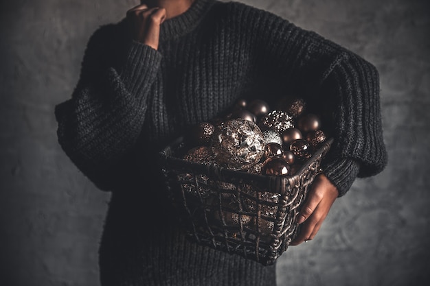 Person in dark sweater holds a basket of bronze, silver, and glittery Christmas ornaments against a moody background.