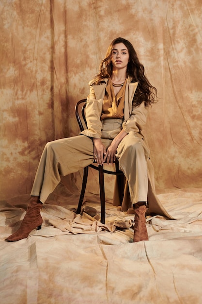 Stylish woman in beige trench coat, brown blouse, wide-leg pants, and matching boots, seated against an earthy brown fabric background.
