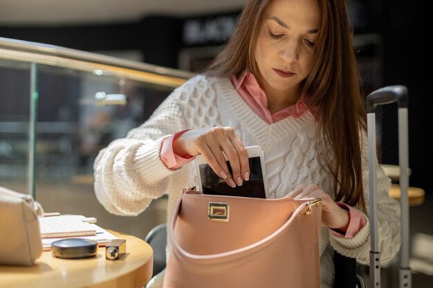 Young woman organizing her pink handbag, placing a white tablet inside. She's wearing a white cable-knit sweater, with luggage nearby.