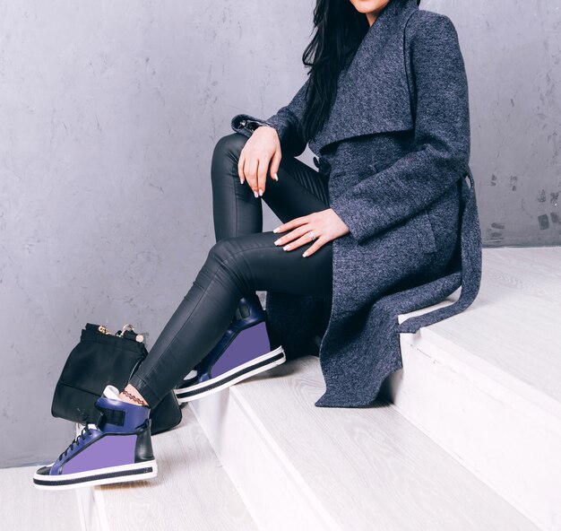 Stylish woman in a long grey coat, black leather pants, and vibrant purple high-top wedge sneakers sitting on white stairs.