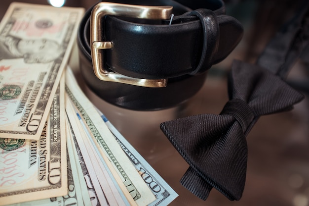 A black leather belt with a gold buckle, a black bow tie, and fanned US dollar bills ($10, $20, $100) on a reflective surface.