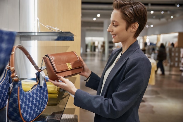 A smiling woman in a blazer holds and examines a brown leather handbag in a bright retail store, with other bags on display.