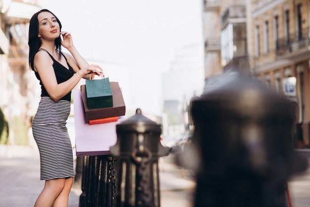 Stylish woman in a striped skirt and black top holding colorful shopping bags while talking on her phone on a city street.