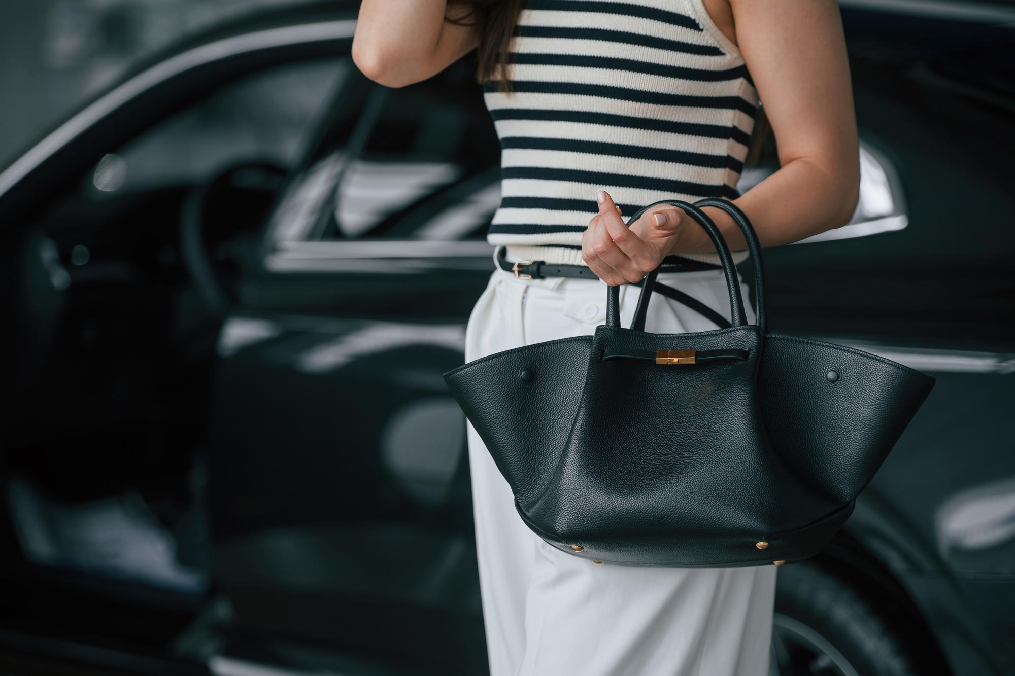 Close-up of a stylish woman in a striped top and white pants holding a black leather tote bag with gold hardware next to a dark car.