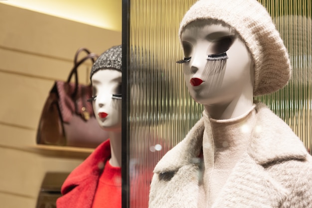Mannequins in a shop window, one in a cream beret and coat, reflected mannequin in a red coat, featuring long lashes.