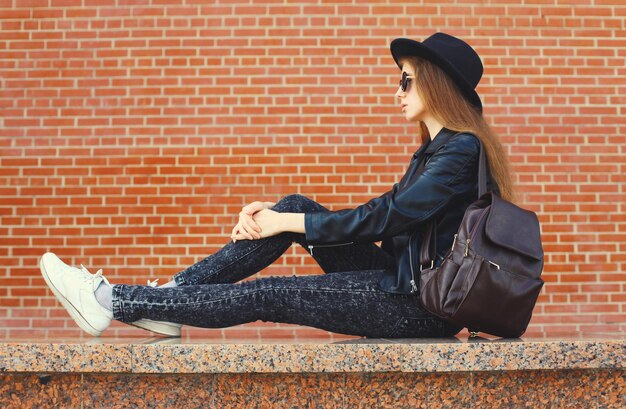 Stylish woman in black fedora, sunglasses, leather jacket, acid-wash jeans, and white sneakers sitting on a stone ledge.