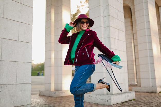 Smiling woman in a burgundy velvet moto jacket, green sweater, cuffed jeans, hat, and sunglasses posing by architectural columns.