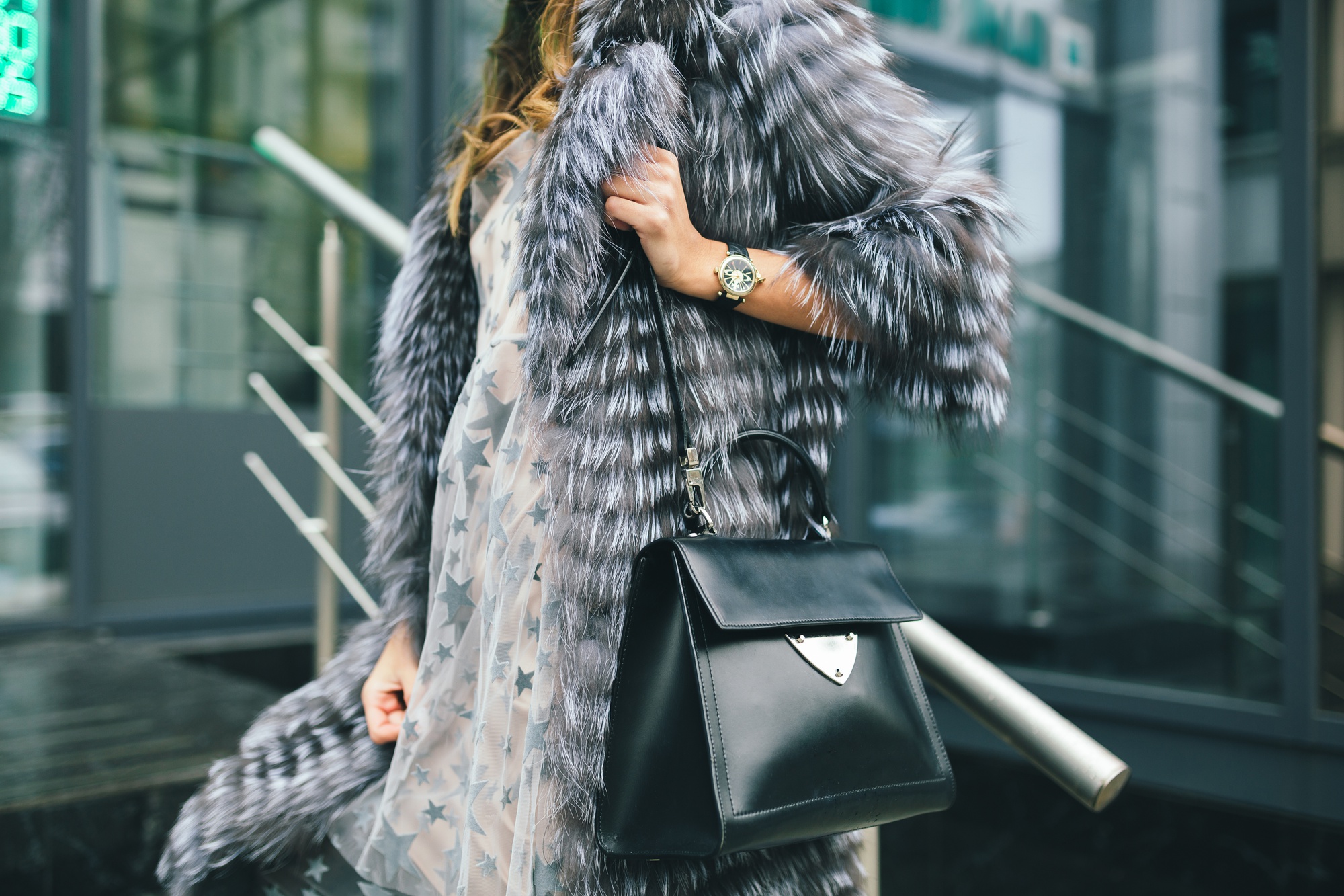 Stylish woman in a luxurious silver fur coat, star-patterned dress, holding a black leather handbag and wearing a gold watch.