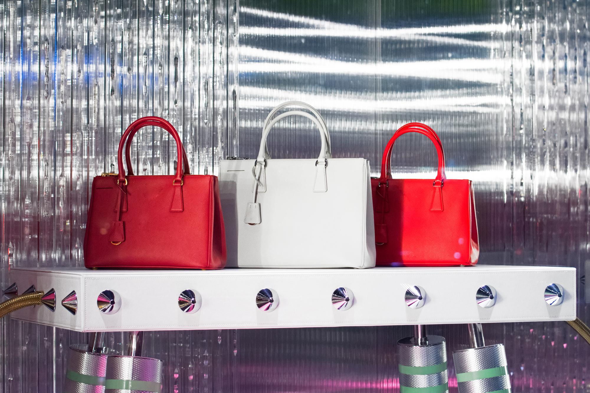 Two red and one white luxury handbags on a sleek white shelf with silver accents, set against a shimmering, reflective background.