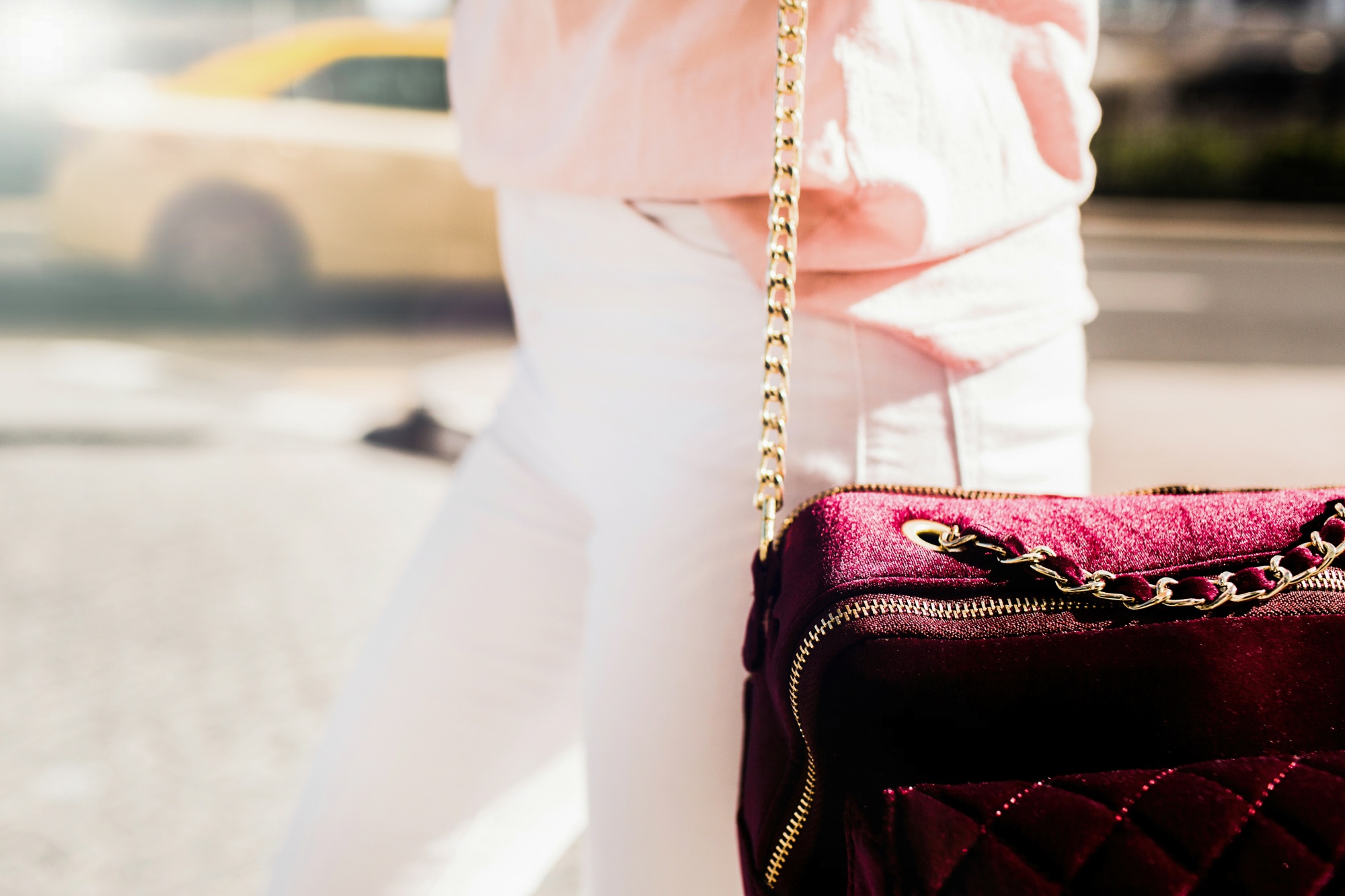 Stylish person in pink and white carries a quilted burgundy velvet handbag with a gold chain strap on a city street.