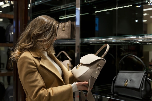 Woman in camel coat examines a light beige handbag in a luxury store, other designer bags displayed in background.