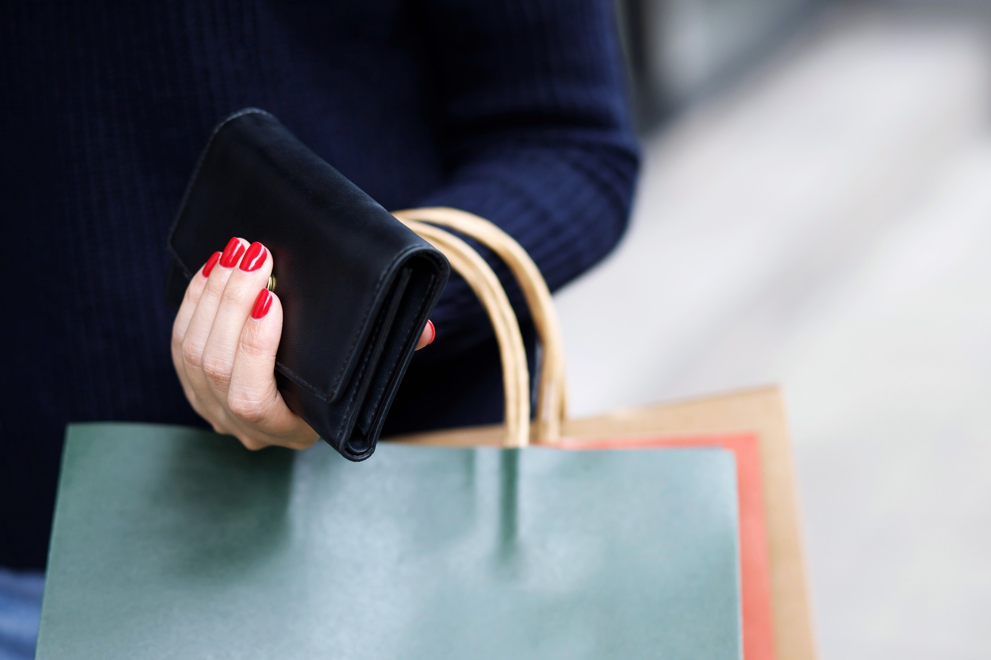 Woman's hand with red nail polish holding a black wallet and shopping bags, ready for a purchase.